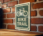 A green and white sign with a bicycle symbol is mounted on a brick wall, indicating a bike trail.