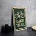 a green and white sign that reads "Bike Trail" with a bicycle symbol on it, placed on a gray countertop next to some black camera lenses.