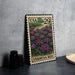 A metal stamp depicting a field of purple flowers, with the text "VERBENA BED" printed on it, is placed on a table next to some black camera lenses.