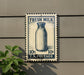 A vintage-style postage stamp featuring a milk bottle, mounted on a wall next to a plant.