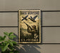 A vintage duck hunting postage stamp is displayed on a wall, featuring a man aiming a rifle at ducks flying overhead.
