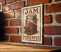 A vintage-style metal sign with the text "JAM PRESERVES" and an illustration of a jar of jam, mounted on a brick wall.