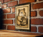 A framed illustration of a hamster holding a small house is displayed on a brick wall.