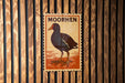 a wooden wall with a vintage-style stamp or postcard featuring a black bird, possibly a moored bird, and the word "MOORHEN" written on it.