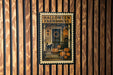 A Halloween-themed metal stamp is displayed on a wooden wall, featuring a porch with a cat, pumpkins, and a wreath.