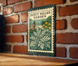 A metal sign with a stamp-like design hangs on a brick wall, featuring a lush garden scene with flowers and greenery.