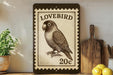 A framed stamp-like artwork depicting a parrot, with the text "LOVEBIRD 20c" printed on it, is displayed on a shelf next to a potted plant and kitchen utensils.