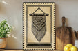 a framed artwork depicting a decorative macrame design hanging on a wall, with a wooden cutting board and a potted plant nearby.