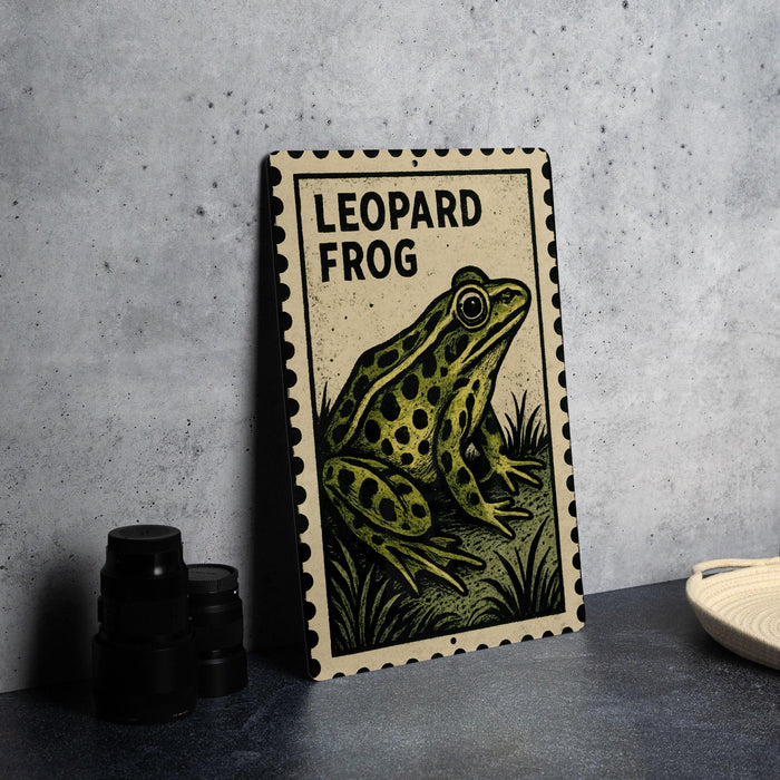 A framed illustration of a leopard frog is displayed on a table, with a stack of black camera lenses and a woven basket nearby.