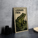 A framed illustration of a leopard frog is displayed on a table, with a stack of black camera lenses and a woven basket nearby.