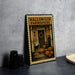 A Halloween-themed metal sign with a porch, pumpkins, and a cat, sitting on a table next to some black objects.