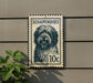 A postage stamp featuring a dog's head is mounted on a wall, with a plant visible to the left.