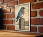A framed illustration of a bluebird perched on a wooden post is displayed on a brick wall.