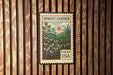 A vintage-style stamp or postcard with a garden scene, featuring plants and a sun, is displayed on a wooden wall.