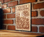a wooden table with a decorative metal sign hanging on a brick wall. The sign is an illustration of a garden scene with various plants and fruits, including a tree, flowers, and squash.