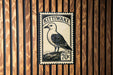 A metal postage stamp featuring a bird, likely a seagull, is displayed on a wooden wall.