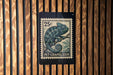 A 25c postage stamp featuring a chameleon is displayed on a wooden wall.
