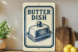 A vintage-style sign that reads "Butter Dish" is displayed on a wooden shelf, accompanied by a wooden cutting board and a potted plant.
