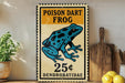 A vintage-style sign advertising a poisonous dart frog for 25 cents, with a blue and black illustration of the frog. The sign is displayed on a wooden surface, with a potted plant and a wooden cutting board visible in the background.