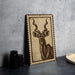 a wooden frame with a detailed engraved illustration of a horned animal, likely a gazelle or similar species, on a gray wall.