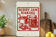 A white sign with a red and black illustration of a jar of jam and a pot of strawberries, along with the text "BERRY JAM MAKING" on a wooden shelf.