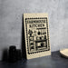 a sign for a "Farmhouse Kitchen" with various kitchen items and utensils depicted on it, placed on a countertop next to some black containers.
