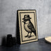 A framed illustration of a bird wearing a hat is displayed on a table, accompanied by a stack of black camera lenses and a woven basket.