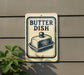 A sign that reads "BUTTER DISH" with an illustration of a butter dish on it, mounted on a wall.