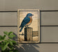 A framed illustration of a bluebird perched on a wooden post, with a plant in the foreground.