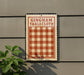 A vintage-style sign with the text "Gingham Tablecloth" is displayed on a gray wall, with a plant visible to the left.