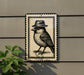 A decorative sign featuring a bird wearing a hat, mounted on a wall next to a plant.