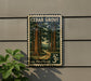 A vintage U.S. postage stamp depicting a scenic forest landscape, mounted on a wall next to a plant.