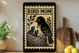 a decorative sign with a bird illustration, placed on a shelf next to a wooden cutting board and some potted plants.