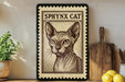 a framed illustration of a Sphynx cat, which is a hairless breed of domestic cat. The cat is depicted in a detailed, monochromatic style, with a wooden frame surrounding the artwork. The framed image is placed on a wooden surface, with a potted plant and a wooden cutting board visible in the background.