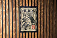 A stamp-like sign with the text "#BIRDS ARE MY FAVORITE PEOPLE" and a portrait of a woman with birds on her shoulder, mounted on a wooden wall.