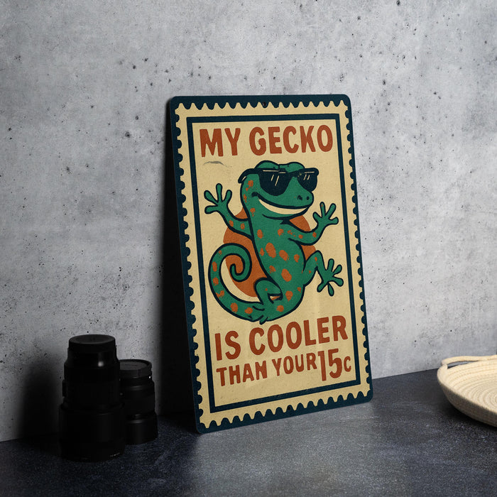 A metal sign with a cartoon gecko wearing sunglasses and the text "MY GECKO IS COOLER THAN YOUR 15¢" is displayed on a table.
