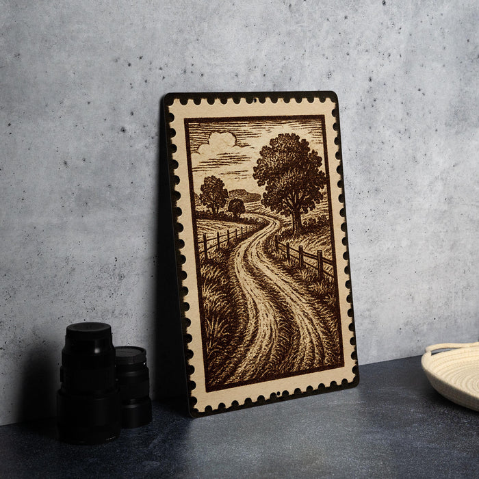 a framed illustration of a rural road with trees and a cloudy sky, placed on a dark surface next to some black camera lenses.