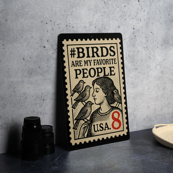 A metal sign with a stamp-like design, featuring a woman and birds, placed on a countertop next to some black camera lenses.