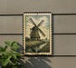 A framed illustration of a windmill is displayed on a wall, with a plant to the left of the frame.