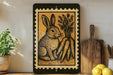 a decorative piece of art depicting a rabbit sitting next to a bunch of carrots, placed on a shelf next to a wooden cutting board and a potted plant.