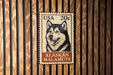 A wooden wall with a framed U.S. postage stamp featuring a black and white image of a husky dog.