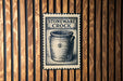 A black and white illustration of a crock pot is displayed on a wooden wall, with the text "STONEWARE CROCK" printed on a postage stamp-like frame.