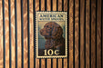 A metal sign with an illustration of a brown curly-haired dog, resembling a stamp, mounted on a wooden wall.