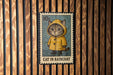 A metal stamp featuring a cat wearing a yellow raincoat, with the text "CAT IN RAINCOAT" printed on it.