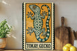 A metal sign featuring a colorful illustration of a gecko, with the text "TOKAY GECKO" below it. The sign is displayed on a shelf next to a wooden cutting board and a potted plant.