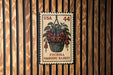 A U.S. postage stamp featuring a hanging basket of fuchsia flowers, mounted on a wooden wall.