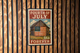 A vintage-style stamp with the text "FOURTH OF JULY USA FOREVER" and an illustration of a barn with an American flag, mounted on a wooden wall.