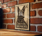 A metal sign featuring a black and white illustration of a dog, with the text "DUTCH SHEPHERD" printed on it. The sign is mounted on a brick wall.