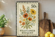 A framed vintage postage stamp depicting a wildflower bouquet, placed on a shelf next to a wooden cutting board and some potted plants.