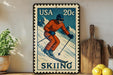 A vintage-style postage stamp featuring a skier in an orange suit and helmet, with the word "SKIING" written below. The stamp is displayed on a shelf next to a wooden cutting board and a potted plant.